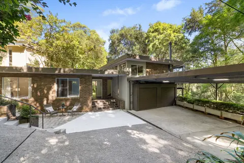 Mid-century brick house with a low skillion roof, brick steps up to an open front door, a paved courtyard with two chairs, and a carport on the right with two roller-door storage bays at the back