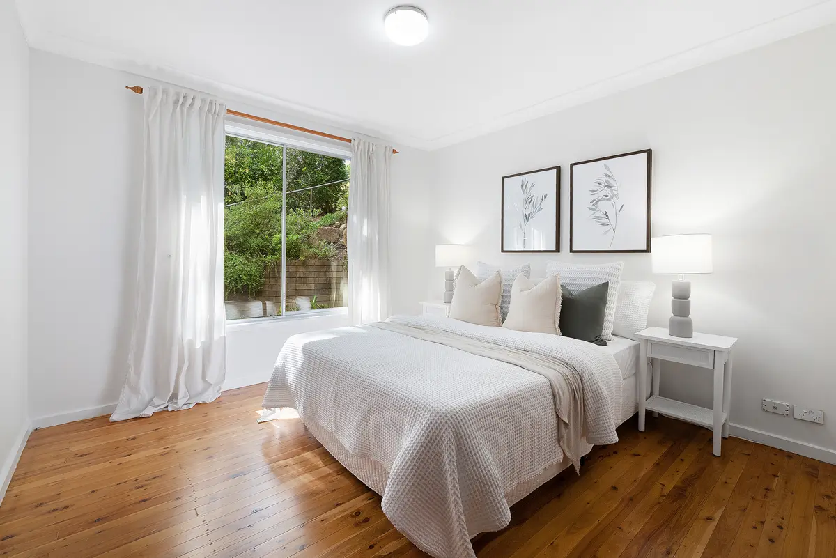 Bright bedroom with a white bed, timber floorboards and a large window overlooking greenery.