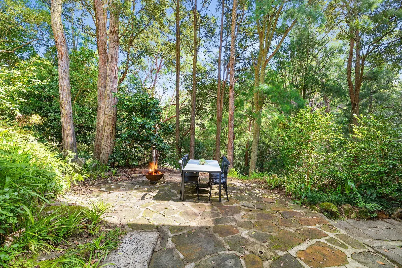 Outdoor table and chairs beside a lit fire pit on a stone patio in a leafy garden.