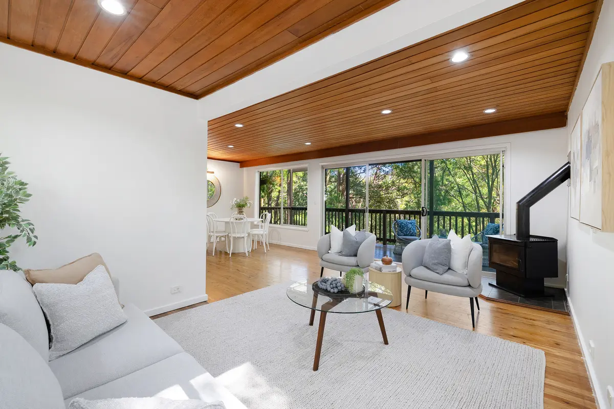 Bright living and dining area with timber ceilings, seating, a wood heater and views to trees through glass doors.