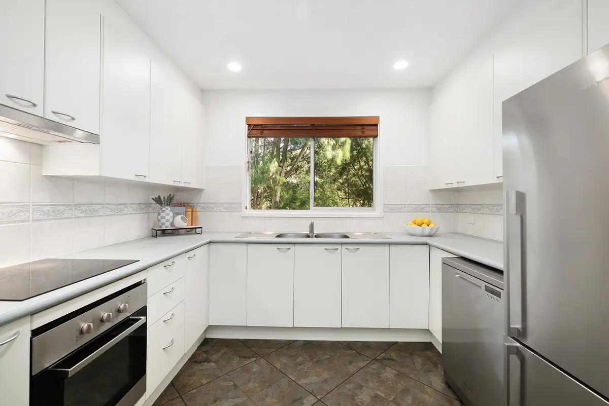 White kitchen with wraparound counters, cooktop, oven, sink, dishwasher and stainless steel fridge beneath a window overlooking trees.